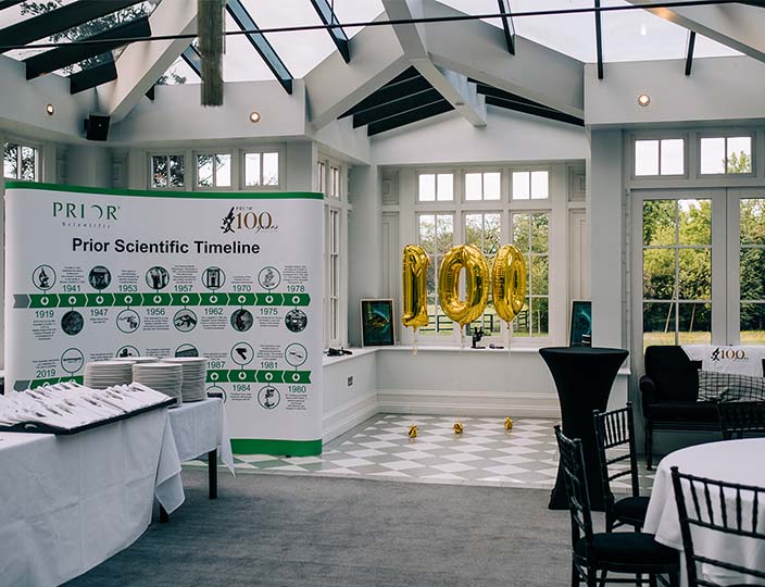 A room set out for a celebration with 100 in helium balloons and a poster showing the timeline of Prior Scientific.